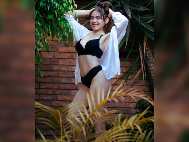 LindaPerez stands with arms up, wearing black bra and panties, white shirt open, showing her tits and ass, smiling, surrounded by green plants and a brick wall.