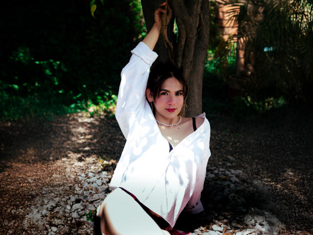 LindaPerez with her tits slightly visible under a white shirt, arms raised, holding tree branches, wearing a pearl necklace, sitting on gravel, dark hair, pink lipstick, sunny garden.