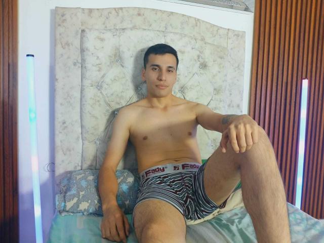 SexyCaicer, shirtless, wearing striped boxer briefs, sitting on a bed with one leg up, looking at the camera, wood panel walls, neon lights, tattoo on right arm.