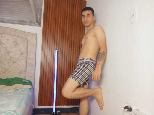 SexyCaicer, shirtless, standing with one leg raised, wearing striped boxer shorts, has tattoos on his right arm, short dark hair, light skin, and a slight smile.