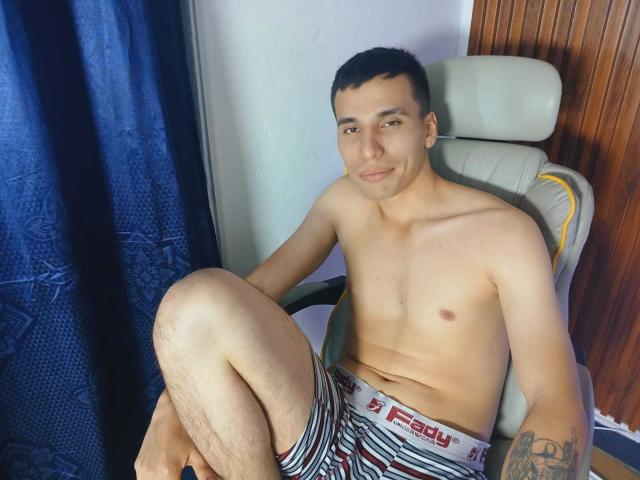 SexyCaicer, shirtless, with short black hair, sitting in a chair, wearing striped red and white "Reddy" boxer shorts, smiling, tattoo on right forearm, legs crossed, wood panel wall.