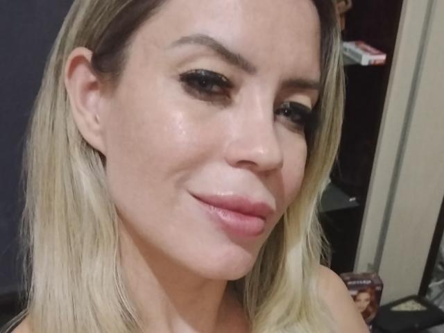 DreamyWomanX with blonde hair, full pink lips, slightly glossy skin, and dark eyeliner, smiling softly at the camera. Her tits are not visible in this image.