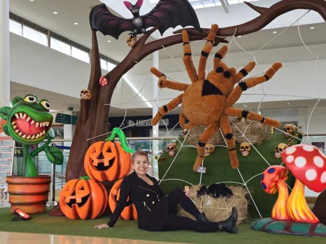 A Halloween-themed setup with LadyLucky sitting in front. She's dressed in a black, form-fitting outfit. The background features a large brown tree with a bat on top and a spider-like structure. Orange pumpkins, a green plant with a wide mouth, and a red mushroom with white spots are scattered around. Hay bales and small skulls add to the spooky decor. LadyLucky has short hair and a neutral expression. Bright indoor lighting.