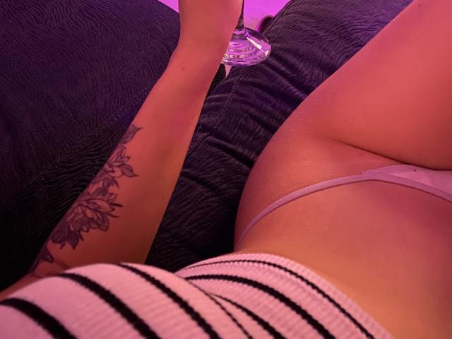 Olieri's getatoë arm houdt een glas vast, haar gestreepte top en witte string laten haar ronde billen en zachte, roze huid zien in paars licht..
