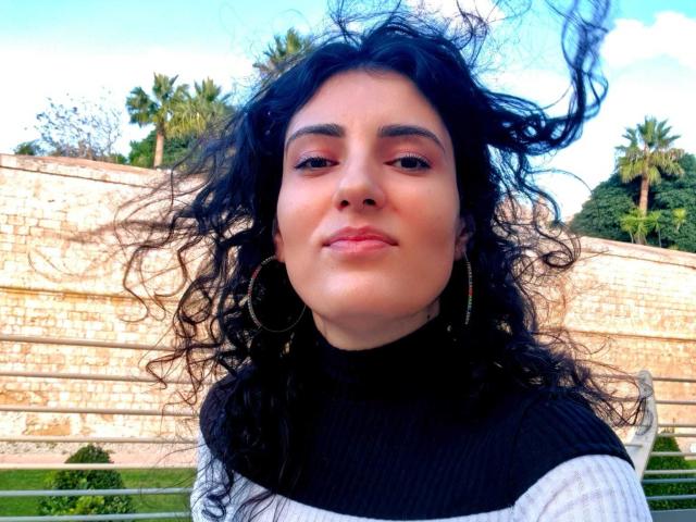 Close-up of AddictedToUTS with medium brown skin, curly black hair, and subtle makeup. She wears a black turtleneck with a white stripe and large hoop earrings. Her hair is windswept, and she looks slightly to the right with a neutral expression. The background has a tall, beige stone wall with greenery and palm trees. Bright daylight enhances the natural colors. The image has a slightly overexposed sky, adding contrast to her face.