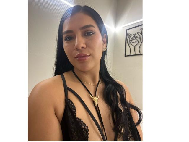 Velka with long black hair, wearing black lace lingerie with strappy design, showing her tits, wearing a gold necklace with a crescent moon pendant, smiling softly.