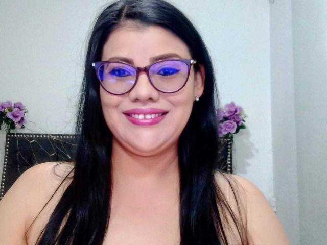 ValeriaLoveer with black hair, purple glasses, pink lipstick, smiling, bare shoulders, purple flowers on either side, white wall background.
