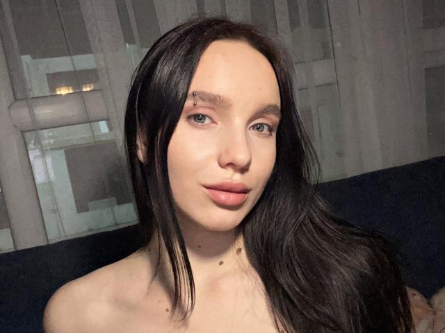 SofiMurr with long black hair, pale skin, and blue eyes, looking at the camera with a slight smile, showing her bare shoulders and a small mole on her neck.