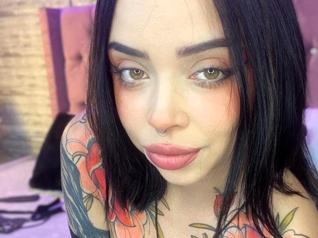NaiaWhisper has black hair, green eyes, a nose ring, and full pink lips. She's topless with colorful tattoos on her chest and arms, looking at the camera.