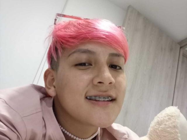 HadeDanny with pink hair, wearing a pink jacket and pearl necklace, has metallic braces and a cute smile, holding a fluffy white toy.