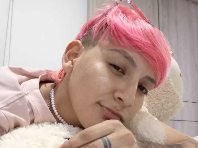 HadeDanny mit rosa Haaren, rasiertem Seitenscheitel, einer Perlenkette, einem rosa Kapuzenpullover, liegt auf einem flauschigen weißen Spielzeug, lächelt leicht, die rechte Hand ruht auf dem Spielzeug, das linke Ohr ist sichtbar..