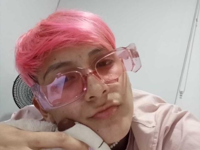 HadeDanny mit rosa Haaren, rosa Brille und einem Nasenpiercing, hält ein weißes Gerät an ihre Wange, trägt eine rosa Jacke..