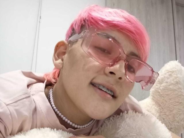 HadeDanny with pink hair, pink glasses, and a pink shirt, wearing a pearl necklace, lying on a white stuffed toy, showing a slightly open mouth with visible teeth.