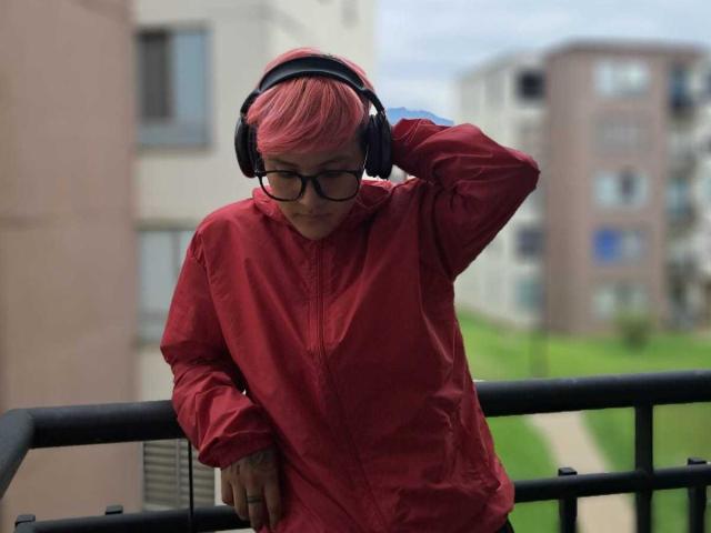 HadeDanny with short pink hair, wearing black glasses and oversized red shirt, leans on a black railing. They're holding a black headphone over their head with their right hand, eyes looking down. Background shows blurred apartment buildings and green grass. HadeDanny's casual pose and muted red shirt contrast with the bright green and grey surroundings. The image has a slightly overcast, blue-grey sky.