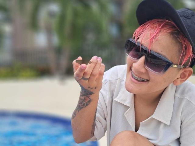 HadeDanny with pink hair, black cap, sunglasses, white shirt, and tattooed hand, smiling near a blue pool, holding a small brown object.