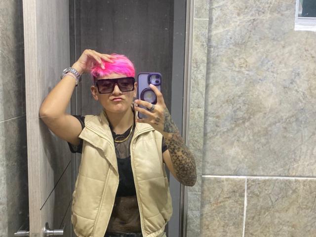 HadeDanny with pink hair, glasses, and tattoos, wearing a beige vest, black shirt, and wristwatch, taking a selfie in a bathroom. Hand on head, phone in hand.