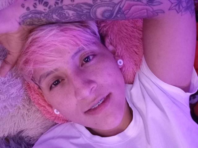 HadeDanny with pink-dyed hair, white shirt, and ear gauges, smiling, arms above head, showing tattoos, lying on pink and white fluffy pillows, under purple light.