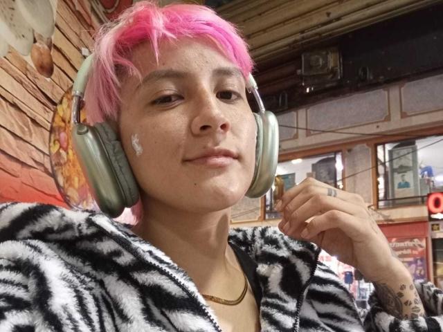 HadeDanny with pink hair, wearing headphones, black-and-white zebra-patterned hoodie, small tattoos on hand, slight smile, light skin, freckles, gold necklace, indoor setting.