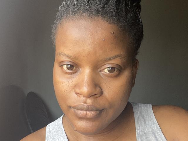 SassyLo has dark brown eyes, short curly black hair, and is wearing a gray tank top. Her brown skin is smooth with small freckles on her nose.