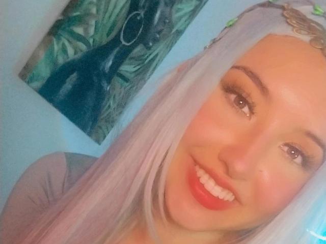 SusanMonroy with long white hair, wearing a green headband, smiling with red lipstick, showing her white teeth, and brown eyes. Her tits are not visible in this shot.