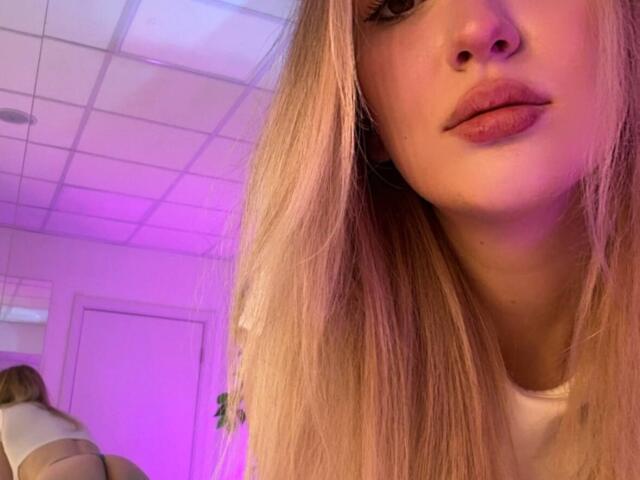 Close-up of Synrix, a blonde woman with long, straight hair and pinkish lipstick. Her face is partially in the frame, with her nose and lips visible. Soft purple and pink lighting illuminates her face from the left. She wears a white shirt. Reflection of her back is visible in the mirror on the left. The ceiling is tiled, and the room has white walls. The image has a casual, slightly blurred quality.