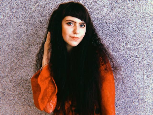 KittyEliza, avec de longs cheveux noirs, une frange, et une chemise orange, la main dans les cheveux, regardant vers le haut avec un léger sourire, peau pâle et yeux bleus..
