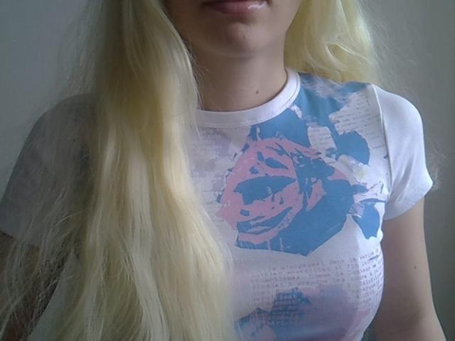 DitaVira has long, wavy blonde hair. She wears a white T-shirt with a pink and blue abstract rose design. The T-shirt has text overlaying the design. Her lips are slightly parted, and her chin is visible. The T-shirt is slightly translucent, revealing the outline of her breasts. The photo focuses on her upper torso and face, with soft lighting and a neutral background.