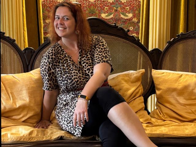 Chleo-hot, red-haired, smiling, wearing leopard print dress, black pants, gold pillows, sitting on ornate wooden couch, yellow curtains, and ornate wall pattern, looking happy.