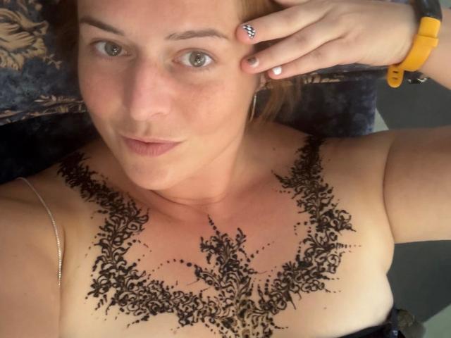 Chleo-hot has a detailed black lace tattoo covering her tits, with light skin and red hair, wearing a yellow wristband, and looking at the camera with a slight smile.