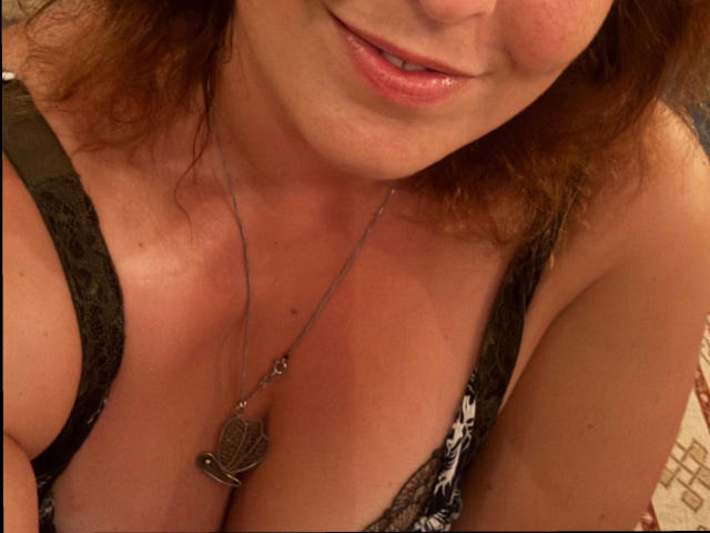 Chleo-hot with her tits showing in a black lace bra, wearing a necklace with a heart-shaped pendant, has slightly parted pink lips and brown wavy hair.