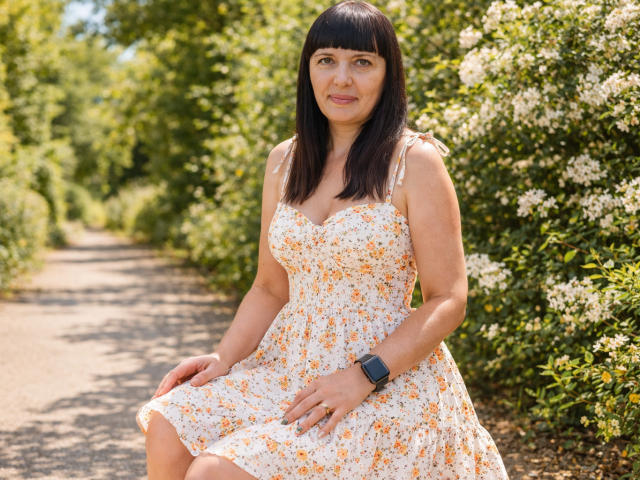 BlairFlepp has medium tits, a cute face, and a floral dress, sitting on a path with greenery and white flowers. She wears a black watch and has straight black hair.