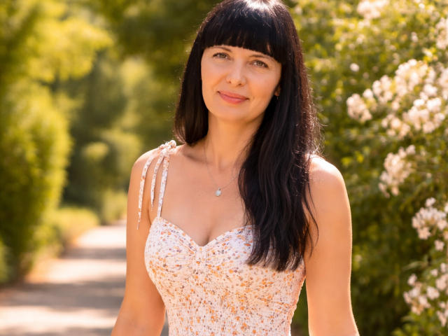 BlairFlepp with black hair, bangs, and a smile, wearing a white floral dress with a sweetheart neckline, small tits, and a necklace, standing on a sunny path.