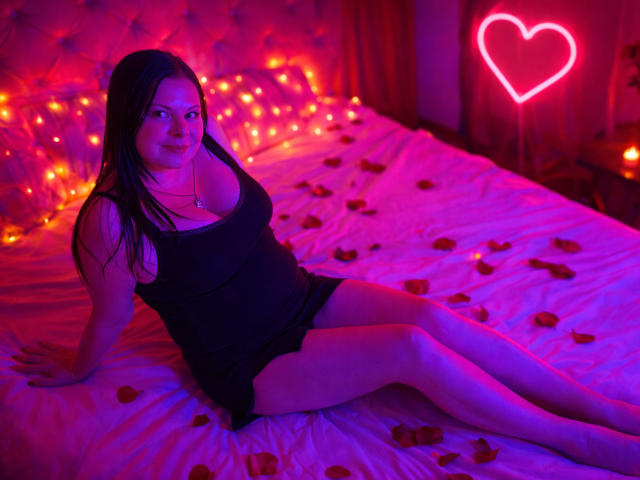 BritneyRaine with long black hair, black tank top, and black shorts, sitting on a bed with rose petals, glowing pink heart neon, and orange fairy lights in the background.