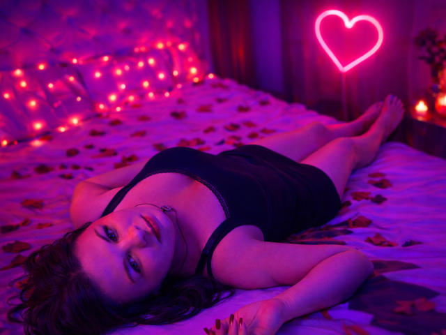 BritneyRaine lies on a bed, illuminated by pink and purple neon lights. She has dark, wavy hair and wears a black, lace-trimmed tank top. Her arms are stretched out, and she looks directly at the camera with a neutral expression. Rose petals are scattered across the bed, and a pink heart neon sign is in the background. BritneyRaine's fair skin contrasts with the dark top and bed. The neon lights create a moody, romantic atmosphere. The image is visually striking, with vibrant colors and a sensual, relaxed composition.