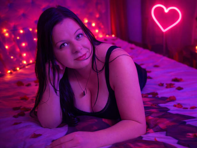 BritneyRaine lies on a bed with purple and pink lighting, wearing a black lace top. Her long black hair is slightly messy, and she looks directly at the camera with a slight smile. A neon pink heart is in the background. Rose petals are scattered on the bed, and warm fairy lights are visible. Her skin is fair, and her eyes are blue. She has a necklace with a small pendant. The composition is intimate and sensual.
