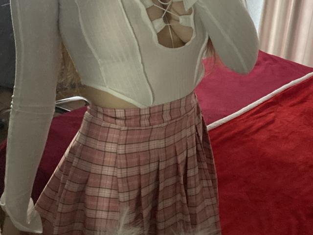 KaiaLein wearing a white, long-sleeve, cut-out top showing her tits, and a pink plaid skirt. Her hand is near her chest. Red bed in the background.