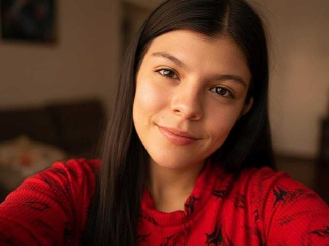 RilleyFoyx a de longs cheveux noirs, des yeux marrons et un sourire discret. Elle porte un pull rouge avec des motifs noirs, et on peut apercevoir une petite tache de rousseur au-dessus de sa lèvre supérieure..