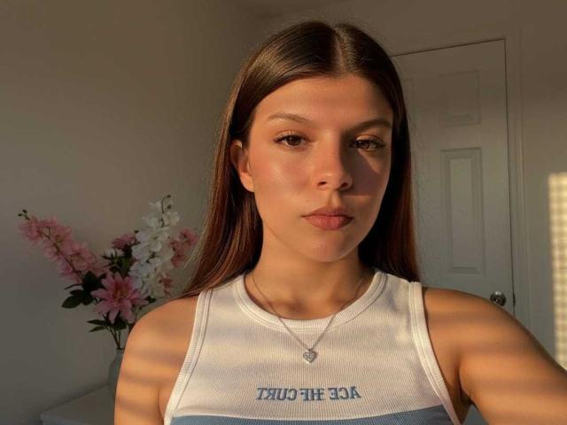 RilleyFoyx with long brown hair, tan skin, and subtle makeup, wearing a white tank top, showing small tits, and a delicate necklace, in soft sunlight.