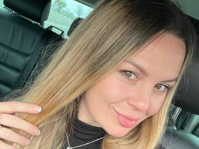 Jilliya with long blonde hair, light skin, and green eyes, smiling slightly, wearing a black turtleneck, and a thin silver necklace, holding her hair with one hand.
