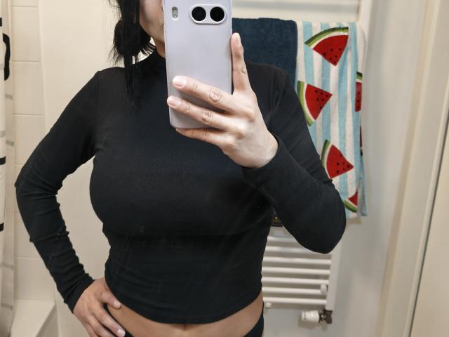 Charlotte Gossart, usando uma cropped preta de manga comprida, mostrando seus seios, segurando um celular, tirando uma selfie no espelho, em um banheiro, com uma toalha de melancia ao fundo..