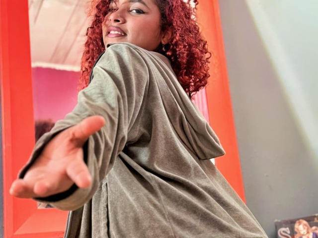 RubyLuna with curly red hair, wearing a green hoodie, reaching out with a finger, smiling at the camera, looking over her shoulder. Her ass is partially visible.