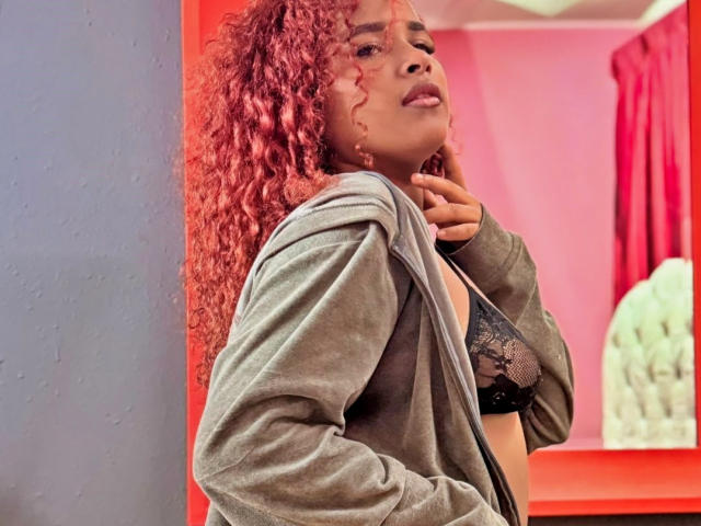 RubyLuna with curly red hair, gray hoodie, and black lace bra, hand near mouth, slight smile, pink wall, red curtains, and green bed in background. Boobs visible through lace.