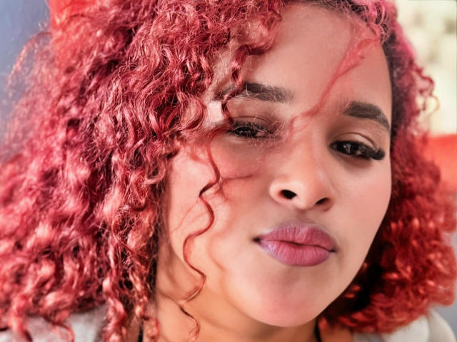 RubyLuna, con i capelli rossi ricci, le labbra rosa piene e le sopracciglia scure, guarda leggermente di lato con un'espressione calma. La sua pelle è marrone chiaro e indossa l'eyeliner..