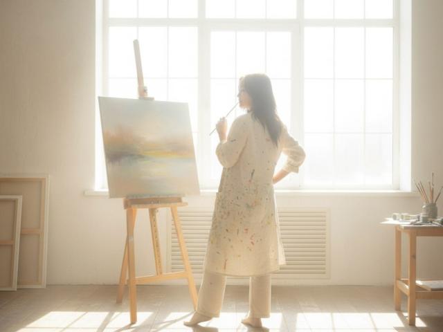 BeloverNana stands in a sunlit room, painting on an easel. She wears a long, white, paint-splattered dress and beige pants, with long dark hair. She holds a paintbrush, looking at the canvas. The canvas is a pastel, abstract landscape with soft colors. The easel is wooden, and BeloverNana is slightly turned to the right. Bright sunlight streams through a large window behind her, casting shadows. A small table with paint supplies is on the right. The room is simple and bright.