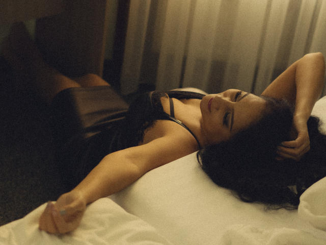 KarinaCecillia lies on a white bed, her dark hair splayed across the pillow. She wears a sheer black dress, arms relaxed above her head. Her eyes are closed, lips slightly parted. The soft, warm lighting casts gentle shadows on her olive skin. Her right arm has a small tattoo. The image has a muted, almost vintage filter, adding a sense of intimacy and calm. The composition emphasizes her relaxed, sensual posture.