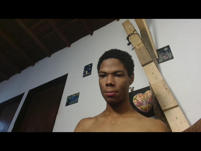 MasterXx is a young Black man with short, curly hair, standing shirtless in a room. He has a medium build, smooth skin, and a serious expression. A small gold necklace is visible around his neck. The background features a wooden ladder to the right and several small, colorful posters on a white wall. The ceiling is wooden, and the room has dark doors. The lighting is natural. MasterXx's facial features are sharp, and his lips are slightly pursed.