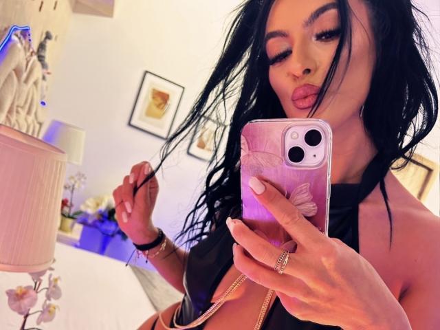 KinkyAmelie with black hair, pouty lips, and a gold ring on her finger, taking a selfie in a mirror, wearing a black halter top, holding a pink phone.