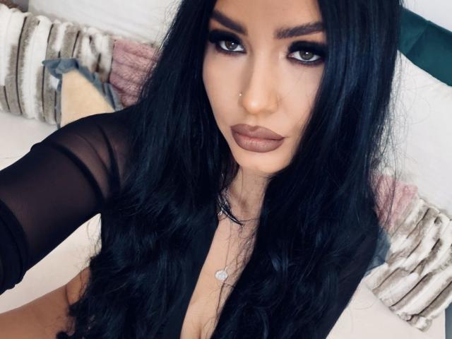 KailynnX with long black hair, dark makeup, and a nose ring, wearing a black sheer top, looking up with full brown lips, small necklace, and a serious expression.