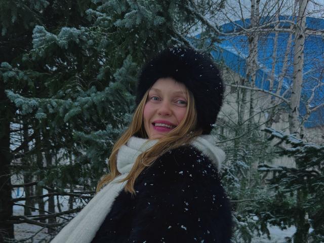 EolantaMarina with long blonde hair, black furry hat, white scarf, black coat, pink lipstick, smiling, snowflakes on her, standing in front of green pine trees, winter.