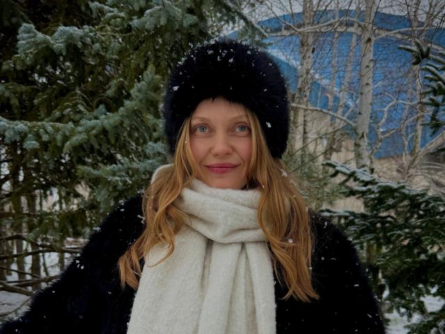 EolantaMarina with long blonde hair, blue eyes, black fur hat, white scarf, black fur coat, snowflakes on face, standing in front of green pine trees, smiling.